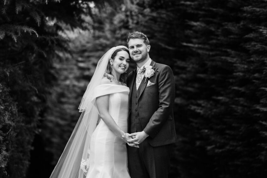 Black and white portrait of the bride and groom holding hands, photographed outdoors with a softly blurred woodland backdrop.