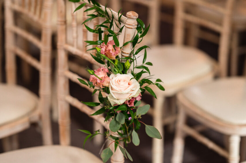 Floral chair decoration with blush roses and greenery tied to wooden ceremony chairs in soft window light.
