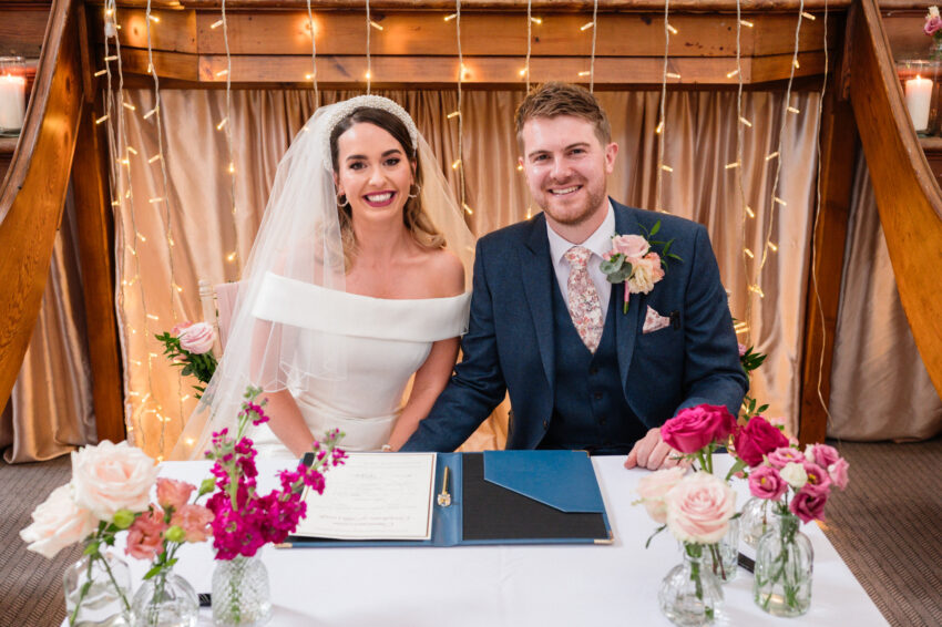 Bride and groom signing the wedding register, seated together beneath warm lights and wooden details at Kings Chapel in Amersham.