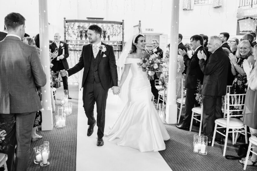 Bride and groom walking back down the aisle hand in hand after the ceremony, guests standing and applauding inside the chapel.