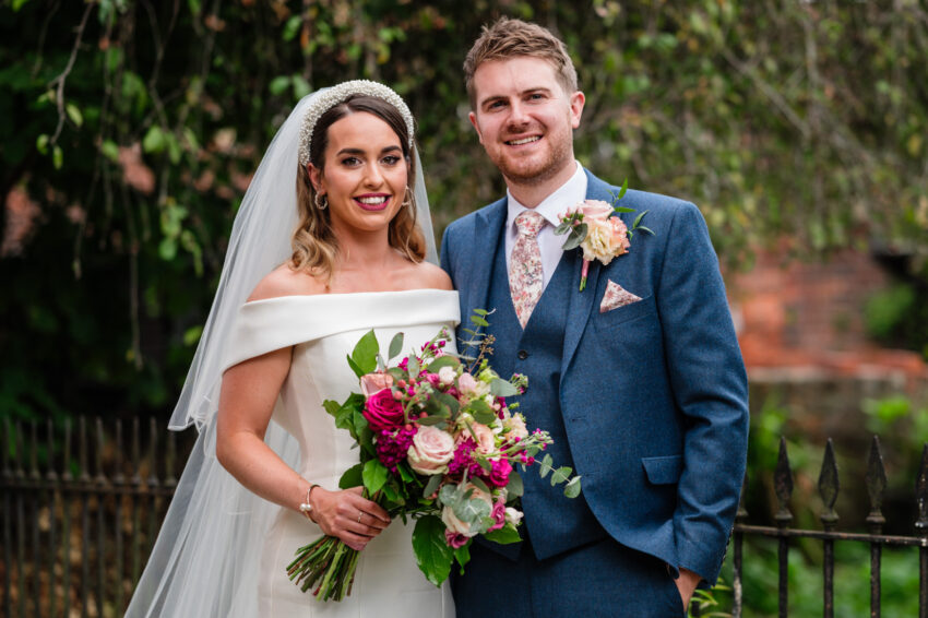 Newly married couple posing together outdoors with bouquets, greenery and soft natural light around them.