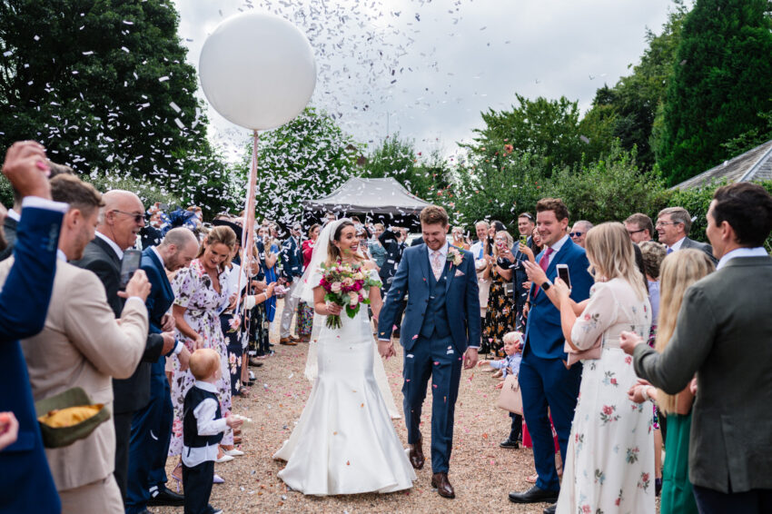 Bride and groom walking through a confetti line outdoors, guests cheering as petals fill the air.