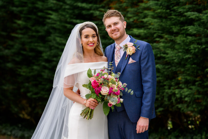 Bride and groom posing together for a relaxed portrait outdoors, framed by soft greenery and evening light.