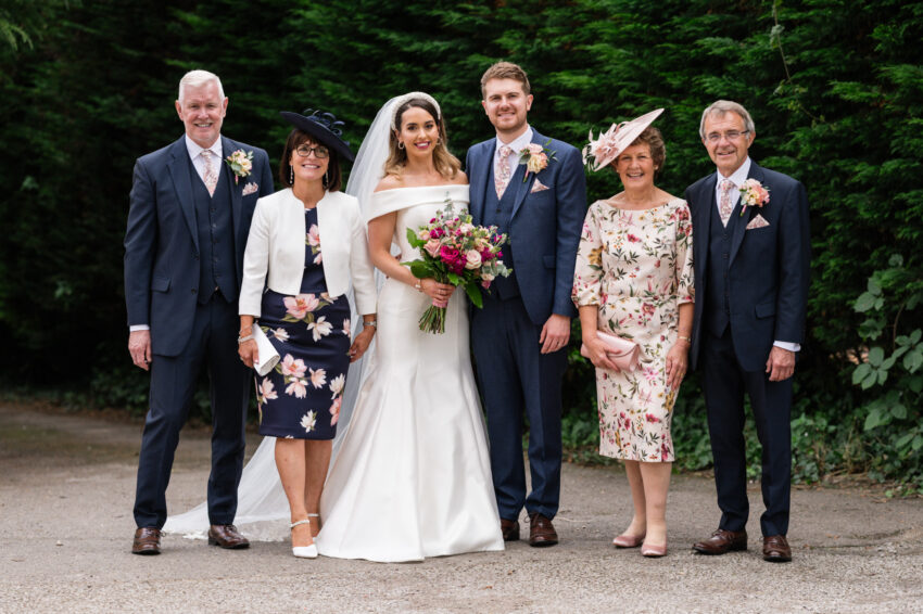 Group portrait of the couple with family members outdoors, formal attire against a leafy backdrop.