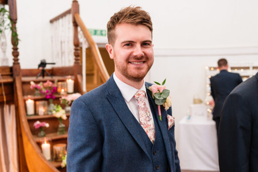 Groom smiling during the ceremony, standing near the front of the chapel with warm candlelight and wooden details behind him.