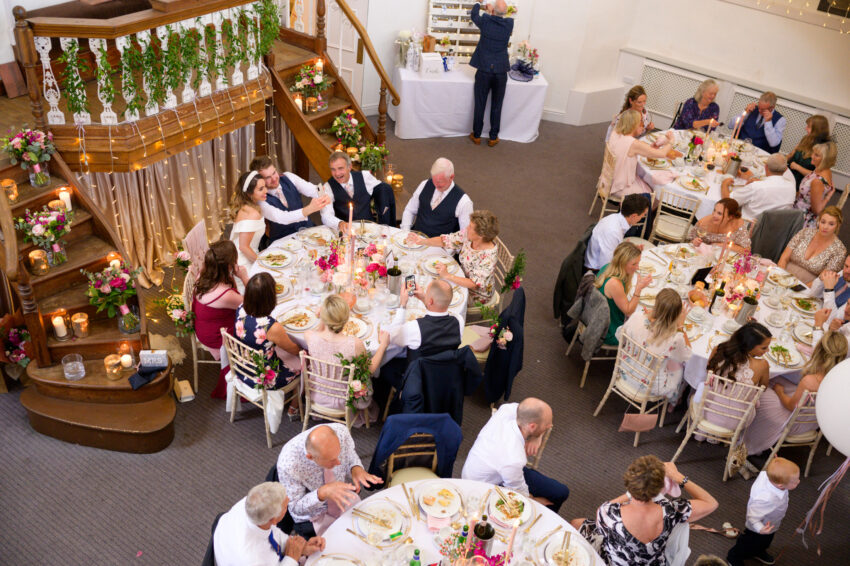 Wedding reception viewed from above, guests seated at round tables beneath warm lights and wooden balconies.