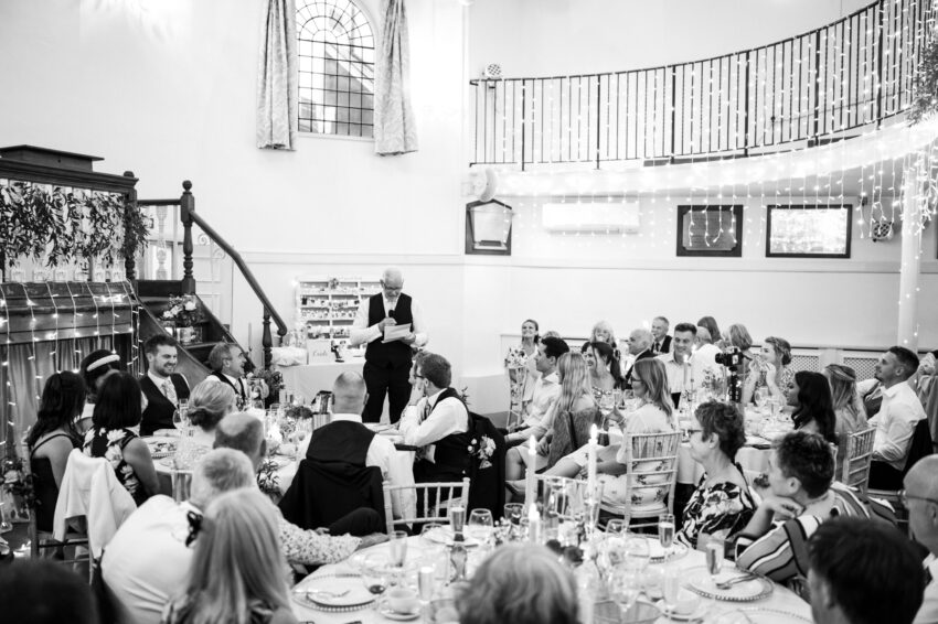 Guests listening to speeches during the wedding breakfast, captured in black and white inside the reception space.
