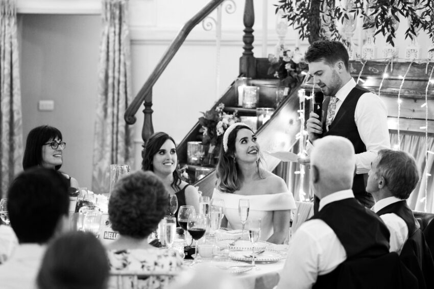 Groom giving a speech during the wedding breakfast, bride and guests smiling at their table beneath soft lights.