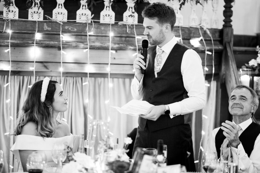 Groom reading from notes during his speech, photographed in black and white with floral table details and hanging lights behind him.