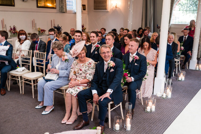 Wedding guests seated inside Kings Chapel in Amersham, watching the ceremony with candles lining the aisle.