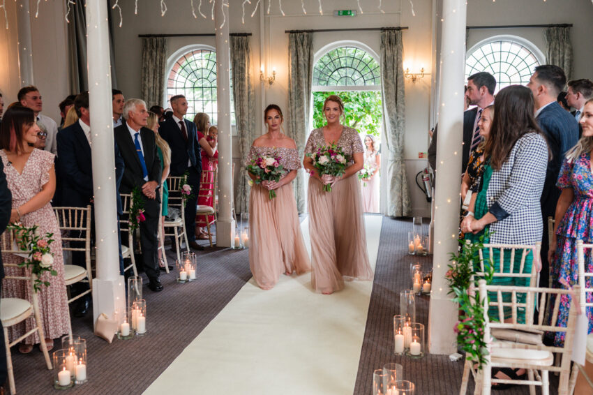 Bridesmaids walking down the aisle in soft pink dresses, carrying bouquets as guests look on inside the chapel.