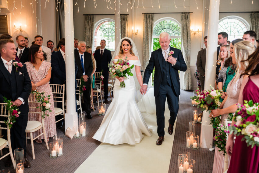 Bride walking down the aisle with her father during a wedding ceremony at Kings Chapel in Amersham, guests watching as candlelight lines the aisle.