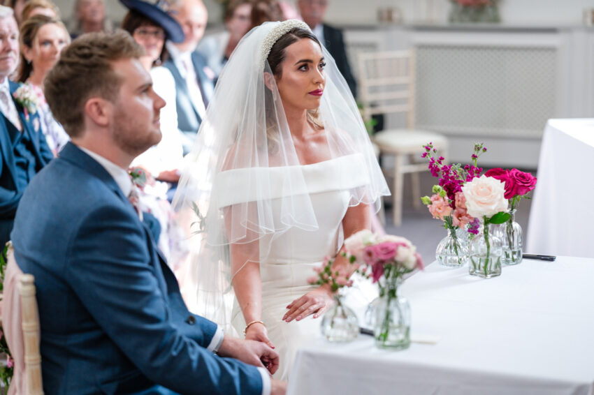 Bride and groom seated together during the ceremony, floral arrangements on the table and guests watching quietly.