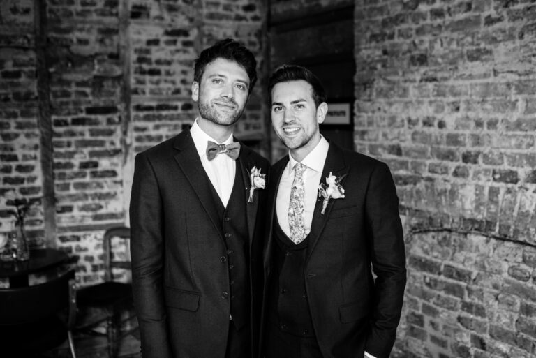 Black and white portrait of two grooms in suits inside a brick wedding venue in London