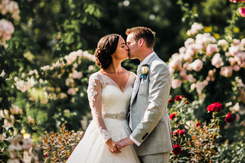 Bride and groom holding hands and kissing in a garden setting, photographed during a quiet moment after the ceremony.