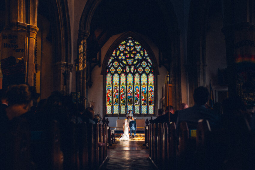 Bride and groom kneeling inside a church, framed by stained glass and soft interior light.