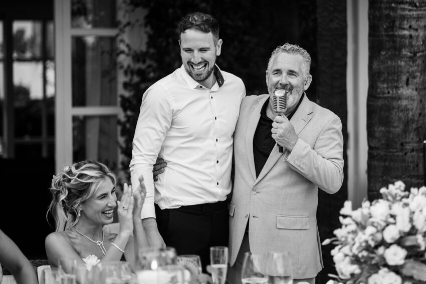 Black and white photo of groom and wedding singer smiling during evening reception at Marbella Club Hotel while bride applauds.