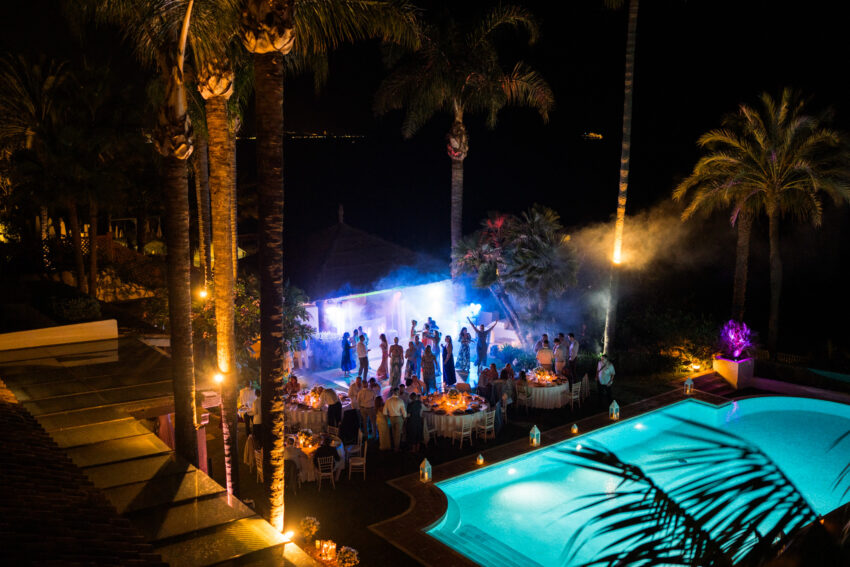 Nighttime view of wedding reception beside the pool at Marbella Club Hotel with guests dancing under palm trees and lights.