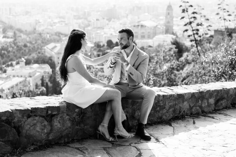Couple sitting together on a stone wall overlooking Málaga in black and white.