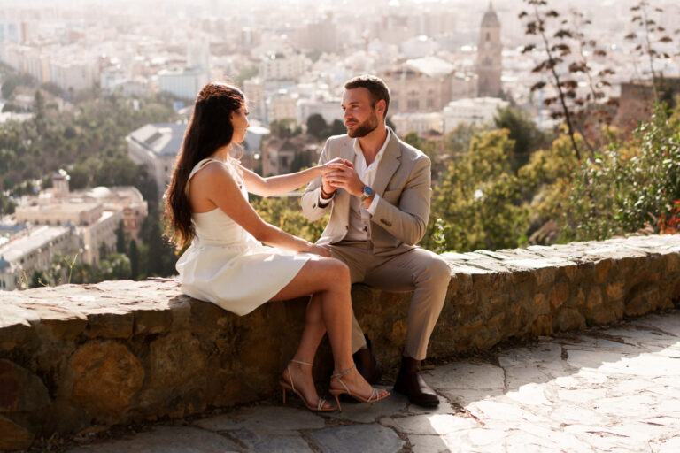 Couple sitting together on a stone wall with views over Málaga during a couples photoshoot.