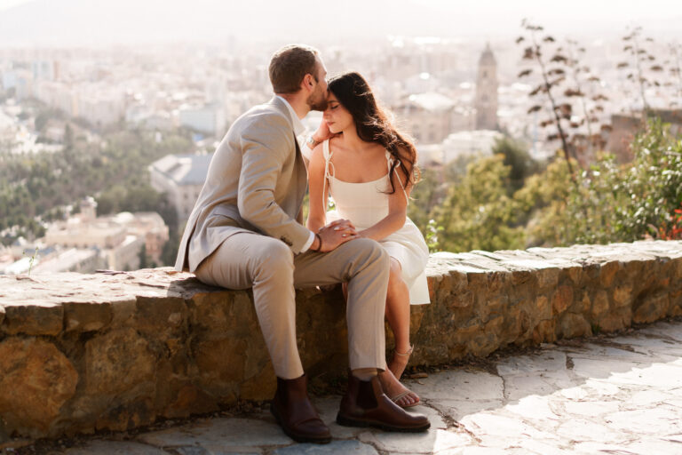 Couple sitting together on a stone wall overlooking the city during a Málaga couples photoshoot on Gibralfaro.