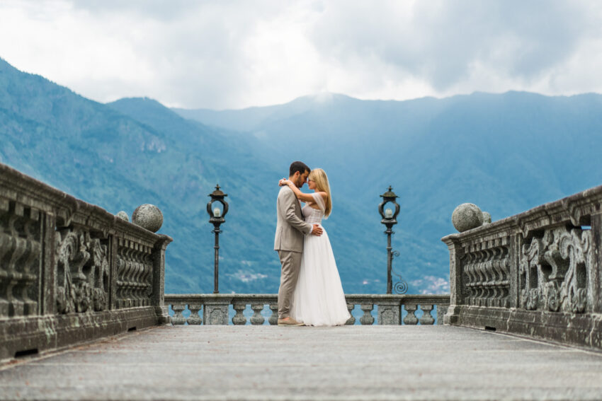 Bride and groom embracing on a stone terrace overlooking Lake Como mountains, romantic editorial wedding portrait