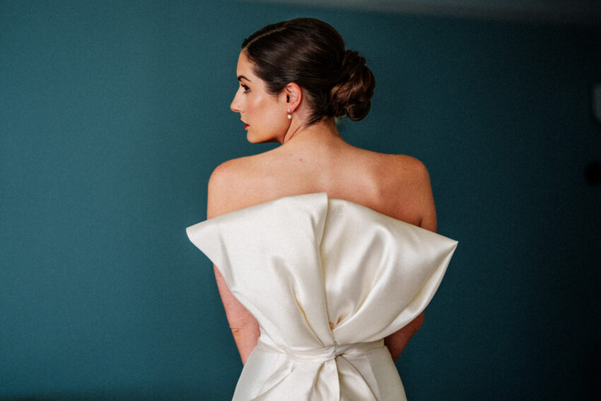 Elegant bridal portrait from behind showing structured wedding dress and low bun hairstyle against deep green background