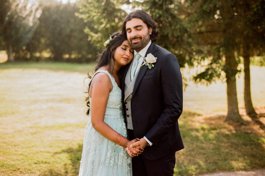 Couple holding hands in golden evening light beneath tall trees, relaxed and intimate portrait with gentle warmth.