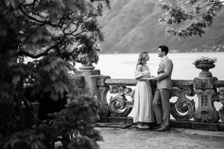 Couple standing at an ornate stone balustrade overlooking Lake Como, framed by foliage in a softly cinematic black and white editorial wedding photograph.