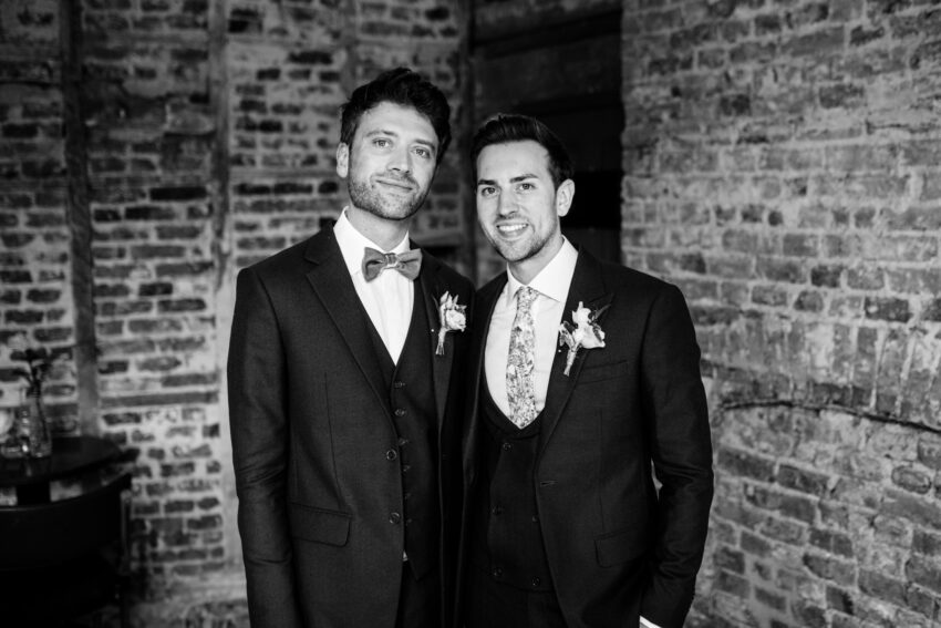 Two grooms in tailored suits against a textured brick wall, relaxed and confident in a modern editorial style portrait.