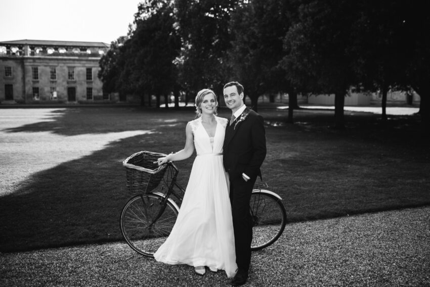 Bride and groom beside a bicycle on a sunlit lawn, timeless black and white portrait with gentle, natural elegance.