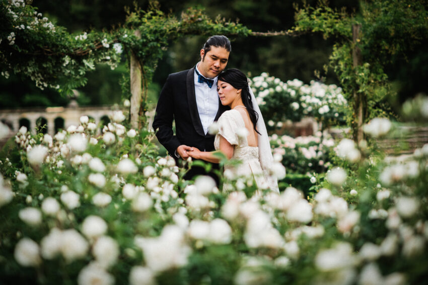 Couple embracing beneath a rose-covered pergola, surrounded by soft white blooms and romantic evening light.