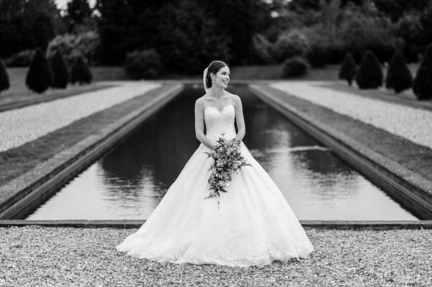 Bride standing beside a long reflecting pool in formal gardens, elegant black and white editorial style portrait