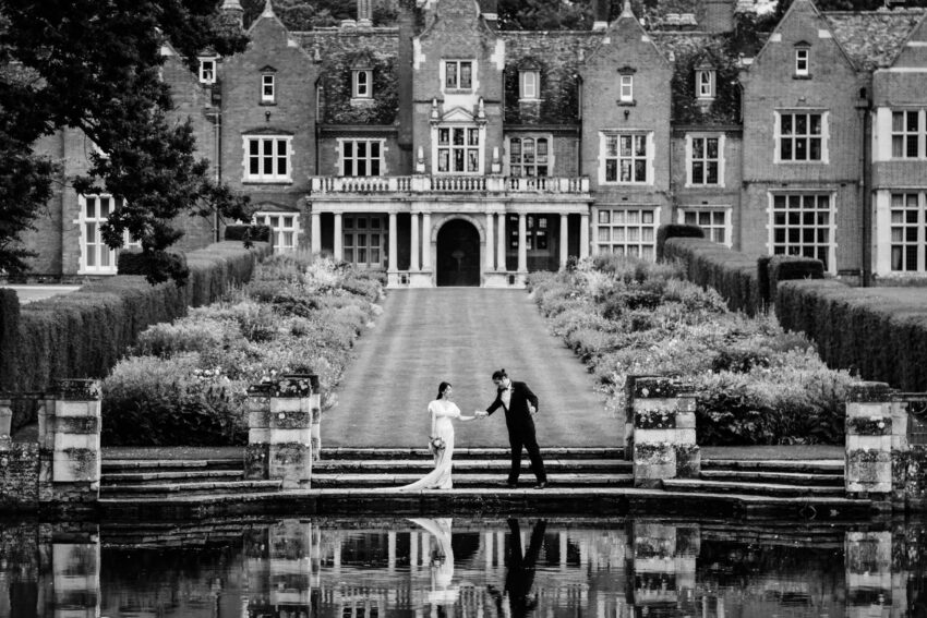 Bride and groom by the lake with Longstowe Hall in the background at Longstowe Hall wedding venue Cambridge.