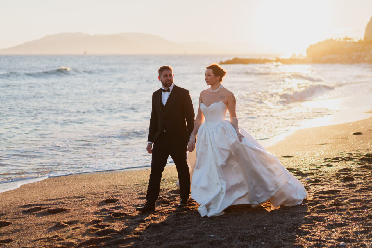 Bride and groom walking along the beach in Malaga at sunset, warm evening light.