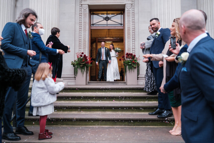 Newly married couple walking out of Old Marylebone Town Hall as guests throw confetti.