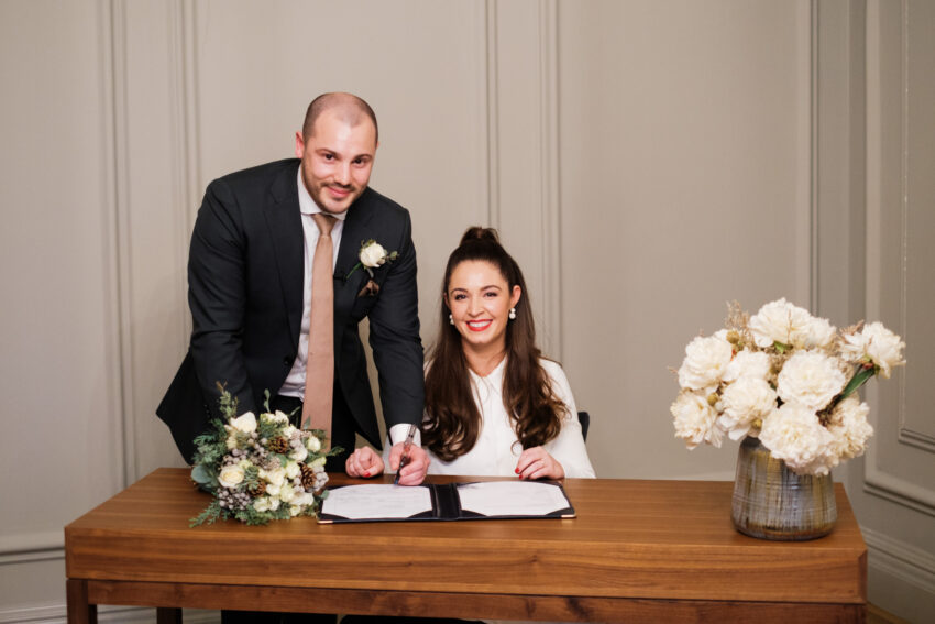 Groom standing beside the bride as she signs the register in the Pimlico Room.