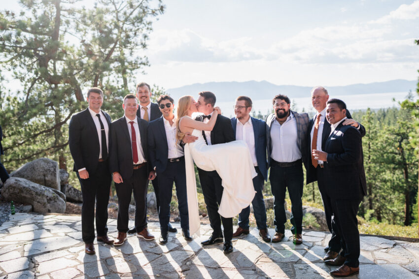 Groom celebrating with friends while holding the bride during portraits overlooking Lake Tahoe.
