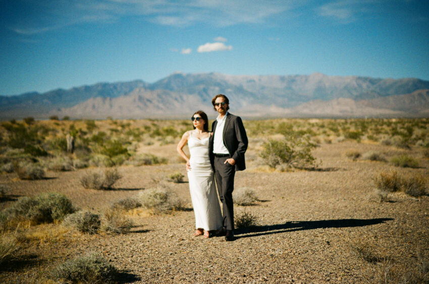 Bride and groom standing together in the Las Vegas desert with mountains in the distance, wearing sunglasses in bright sunlight, captured on 35mm film with soft tones and natural grain, Las Vegas elopement.