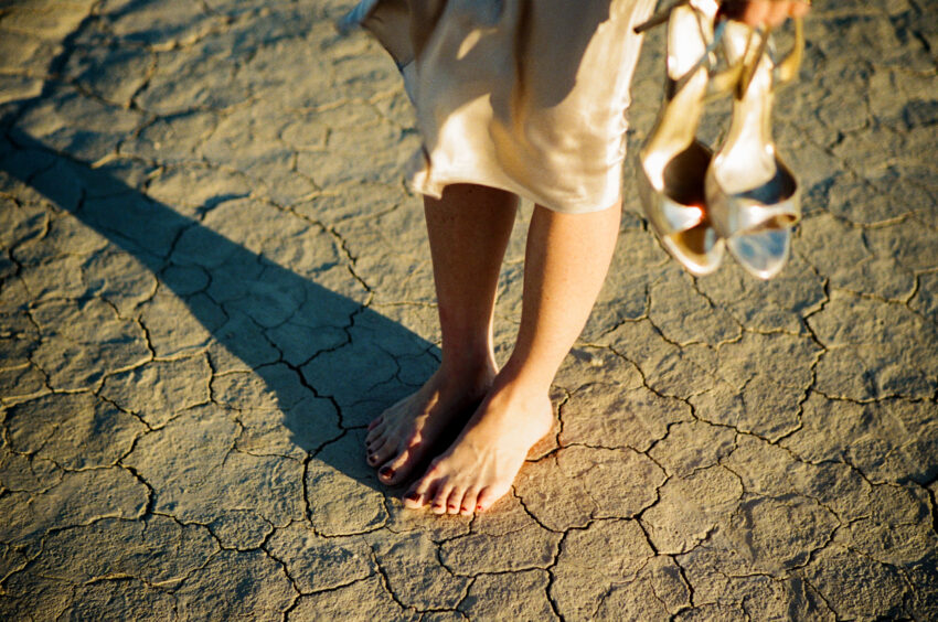 Bride standing barefoot on sunlit dry lakebed, holding wedding shoes, 35mm film.