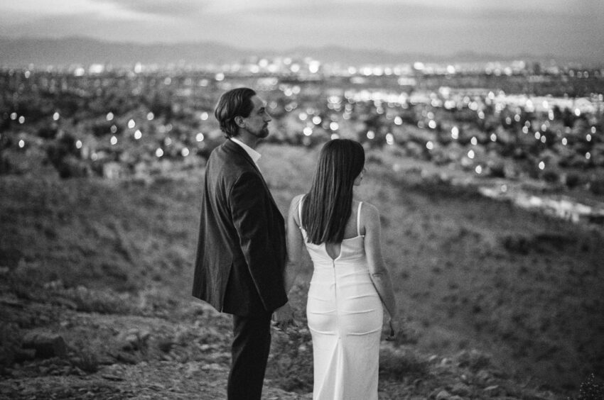 Bride and groom standing hand in hand on a hillside overlooking a city at dusk, city lights softly glowing in the distance, captured in black and white on 35mm film with soft grain and a timeless feel.
