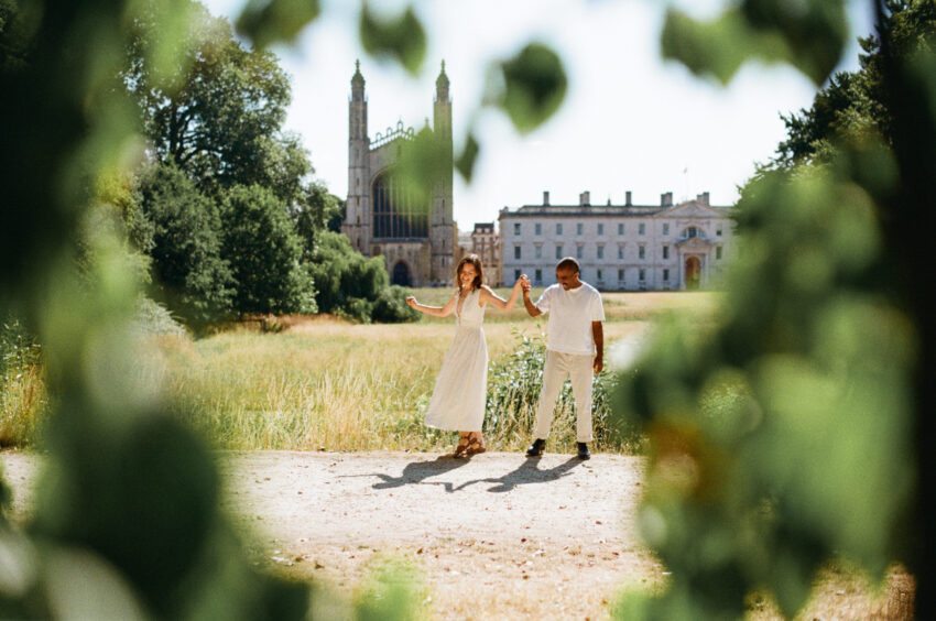 Couple holding hands and dancing through sunlit grass near a historic Cambridge college, framed by soft foreground leaves, captured on 35mm film with natural light and gentle grain.