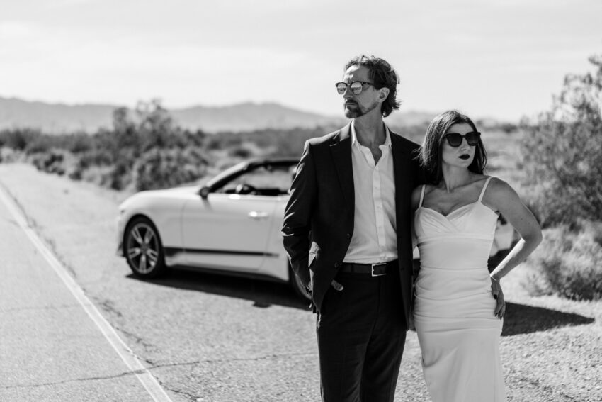 Elegant couple in sunglasses standing beside a convertible on a desert road, photographed in a cinematic black and white style by a Las Vegas elopement photographer.