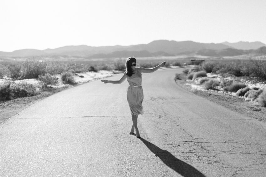 Bride walking barefoot along a quiet desert road with long shadows in black and white, captured in a relaxed editorial style by a Las Vegas elopement photographer.