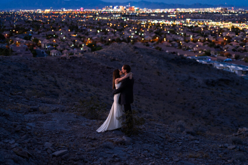 Couple embracing on a hillside overlooking the Las Vegas skyline at night, with city lights from the strip glowing in the distance, photographed by a Las Vegas elopement photographer.