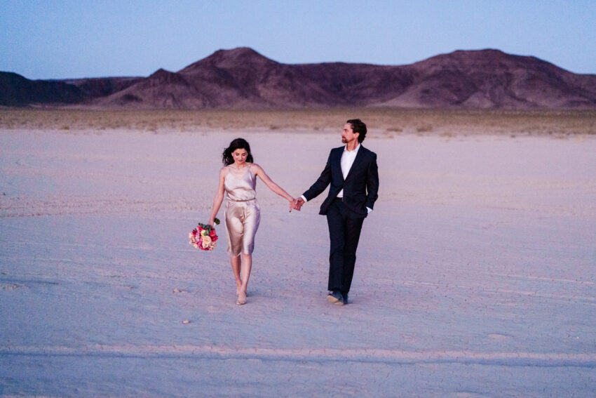 Couple walking hand in hand across a vast desert landscape at dusk, with soft pastel tones and mountains in the distance, photographed by a Las Vegas elopement photographer.