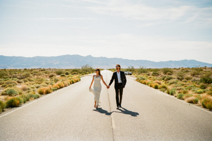 Bride and groom walking hand in hand along an empty road with mountains in the distance, captured on 35mm film with warm tones and natural grain.