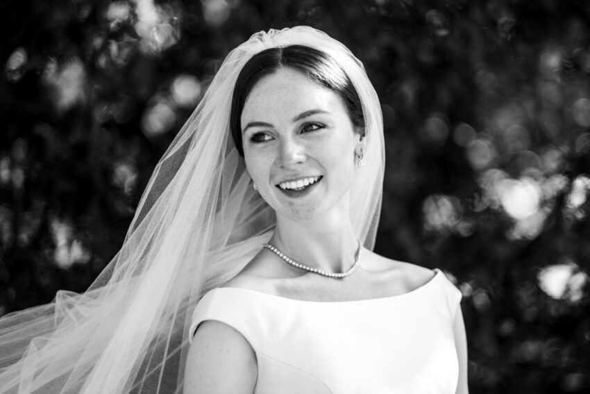 Black-and-white portrait of a bride wearing an elegant off-the-shoulder wedding dress and veil, smiling and looking off to one side. The image is softly lit with a blurred background of foliage, creating a timeless and classic bridal portrait.