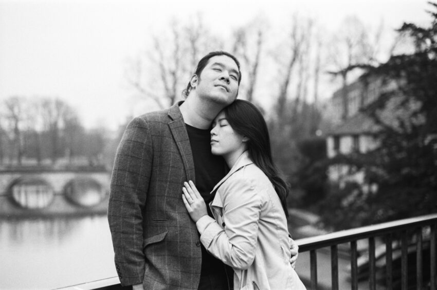 Black and white engagement portrait of a couple embracing beside a river in Cambridge, with soft background blur and bare winter trees. Relaxed, intimate pre-wedding photo with natural emotion and elegant reportage-style composition.