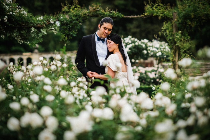 Romantic wedding portrait of a bride and groom standing among white roses in a formal garden, framed by flowers and greenery.